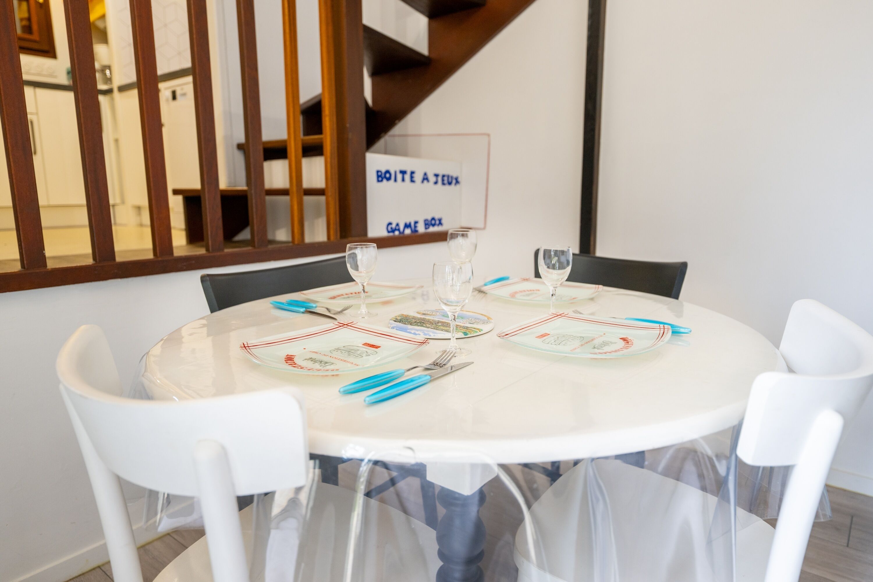 Una mesa redonda para comer con sillas blancas, dispuesta con platos, cubiertos y copas de vino. Un cartel en la pared dice 'BOITE A JEUX GAME BOX'.
