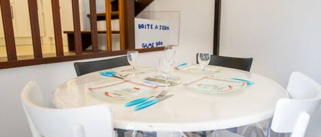 Una mesa redonda para comer con sillas blancas, dispuesta con platos, cubiertos y copas de vino. Un cartel en la pared dice 'BOITE A JEUX GAME BOX'.
