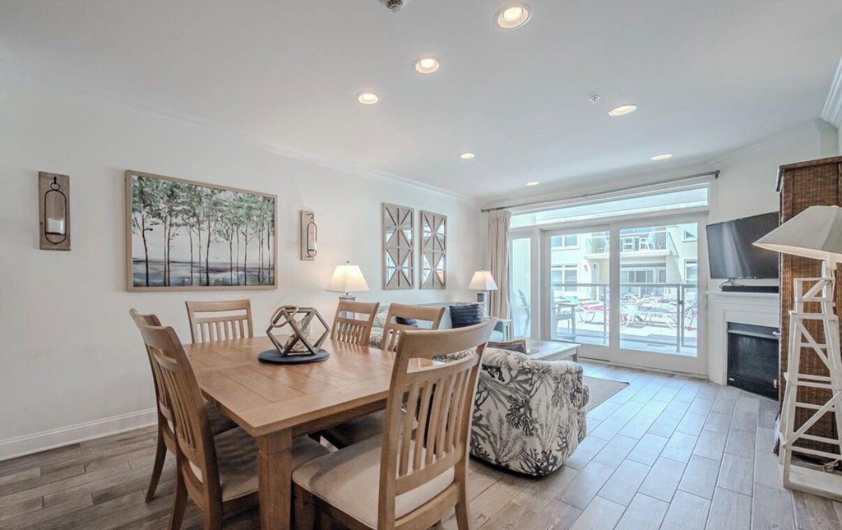 A dining area with a wooden table and chairs, a painting on the wall, and a view through large windows.