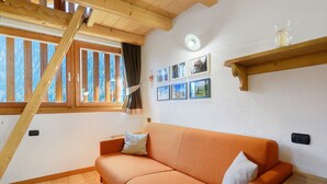 A room with a wooden ceiling, a sofa with cushions, a wooden table, a shelf with pictures, and a window with curtains. - Maso di Zonadi – Alpine House in the Heart of Val di Rabbi (Rabbi)