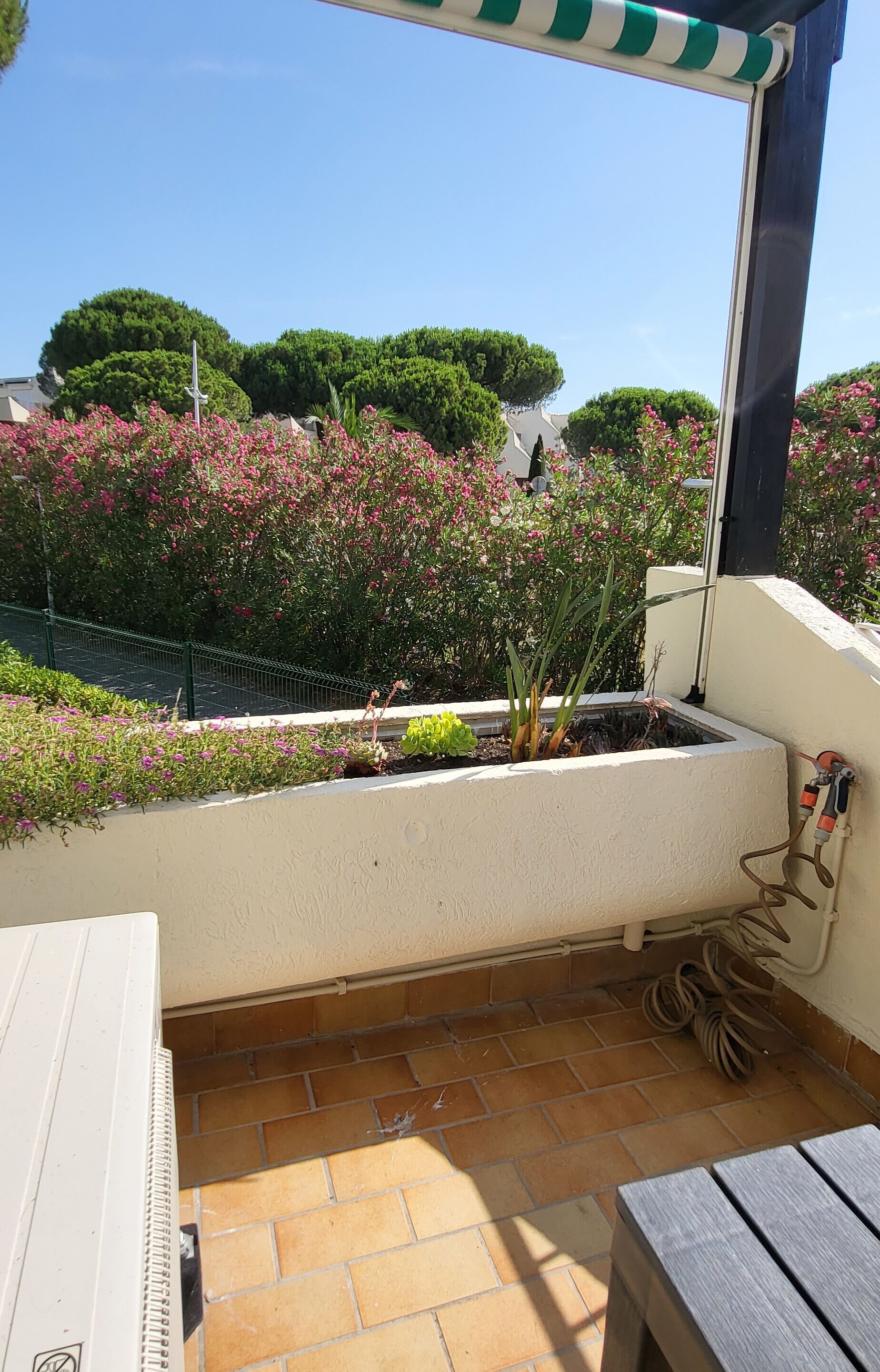 Een balkon met tegels, een tafeltje en een stoel. Achtergrond toont een tuin met roze bloemen en groene struiken.
