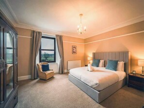 A bedroom with a large bed, a chair, a nightstand, a chandelier, and a window with curtains.