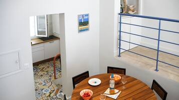 Un coin repas avec une table ronde en bois, des chaises, un saladier de fruits, une bouteille d’eau et un verre à vin. On aperçoit une rambarde bleue et une affiche accrochée au mur.