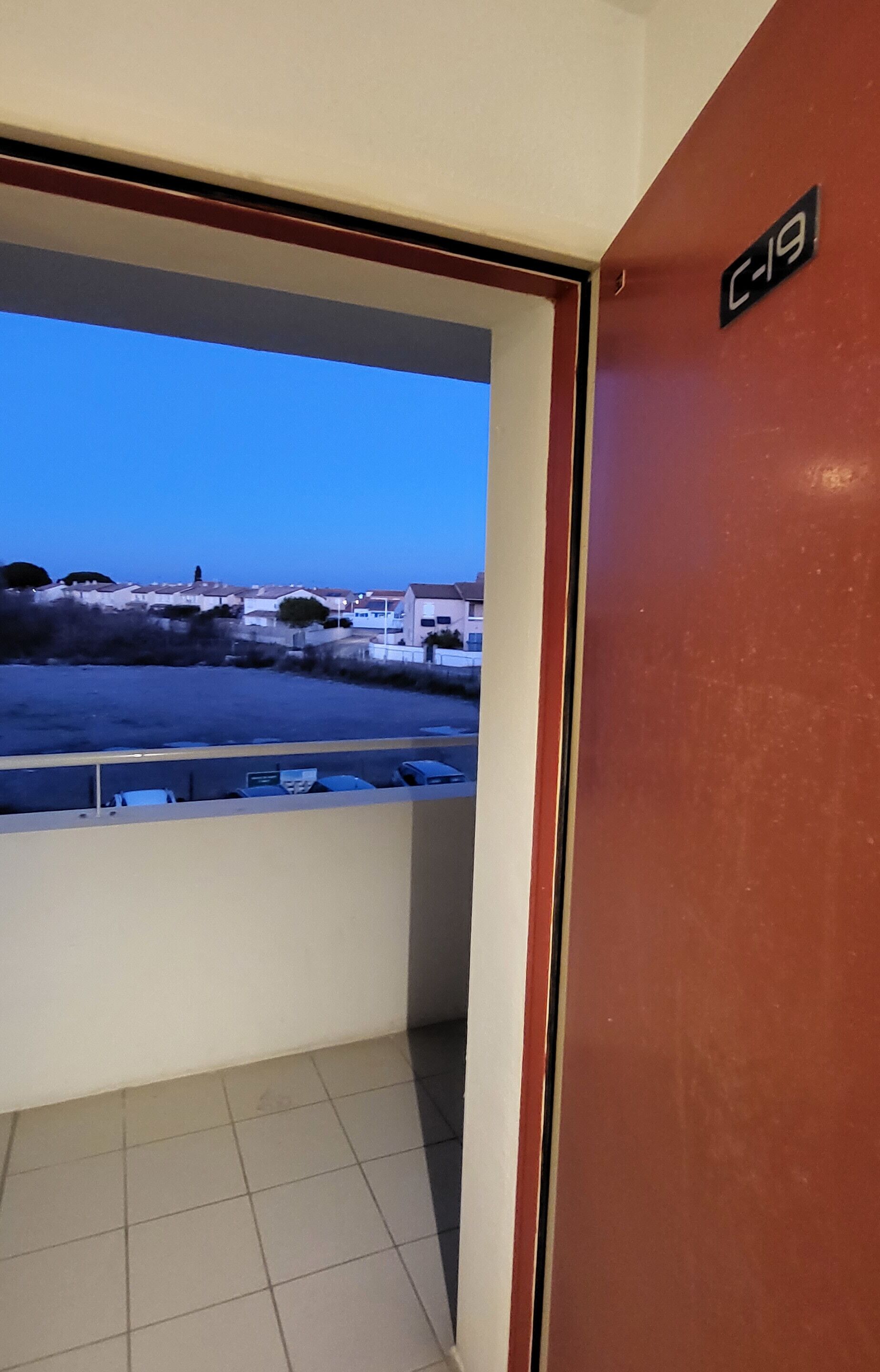 Vista de um quarto com uma porta identificada como “C-19”, em direção a uma área de estacionamento e edifícios ao longe.