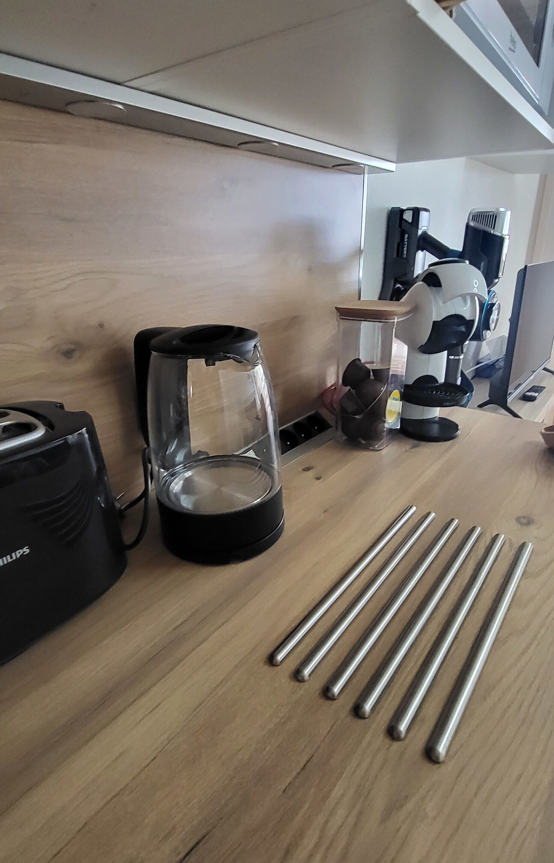 Un comptoir de cuisine avec une machine à café, un récipient en verre doté d’un couvercle et plusieurs tiges métalliques.