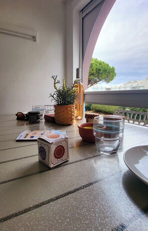 Ein Tisch mit einem Kartenspiel, einem Glas Wasser, einer Flasche und einem Blumentopf auf einem Balkon mit Blick auf Gebäude und Bäume.