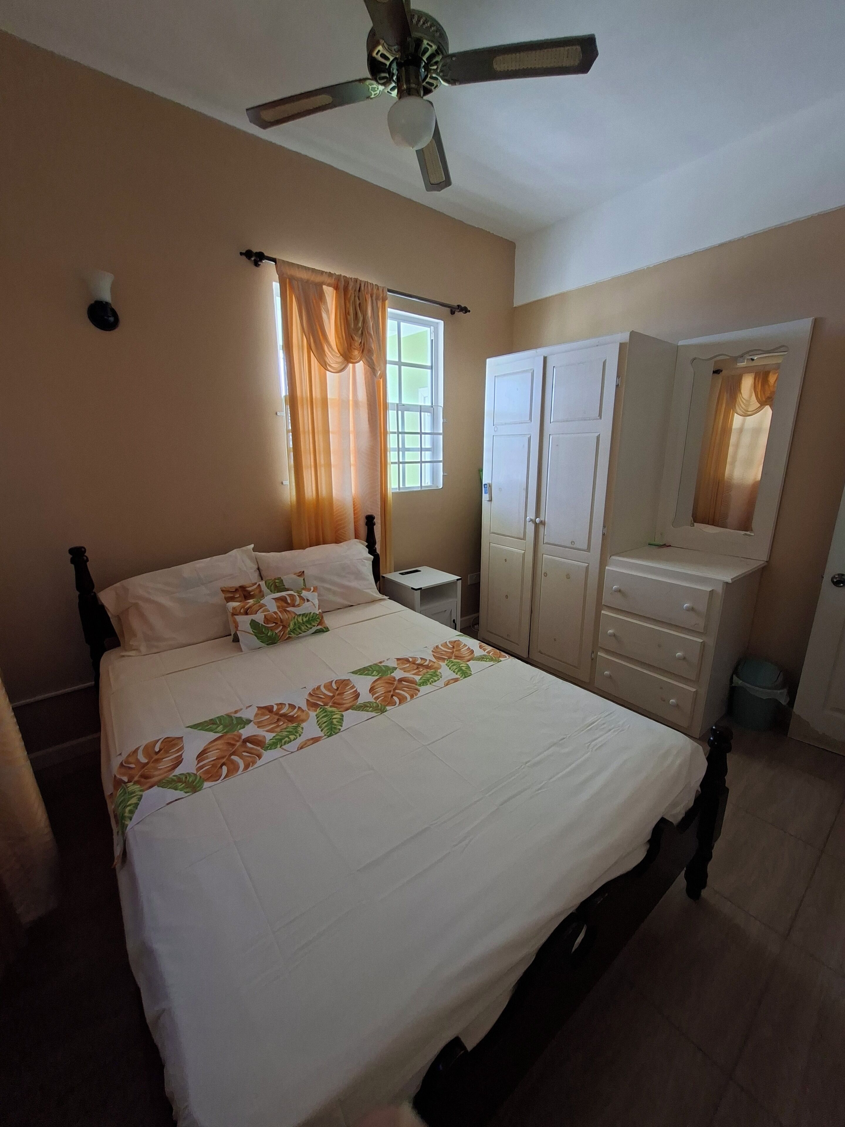A bedroom with a bed, a wardrobe, a mirror, a ceiling fan, and a window with curtains.