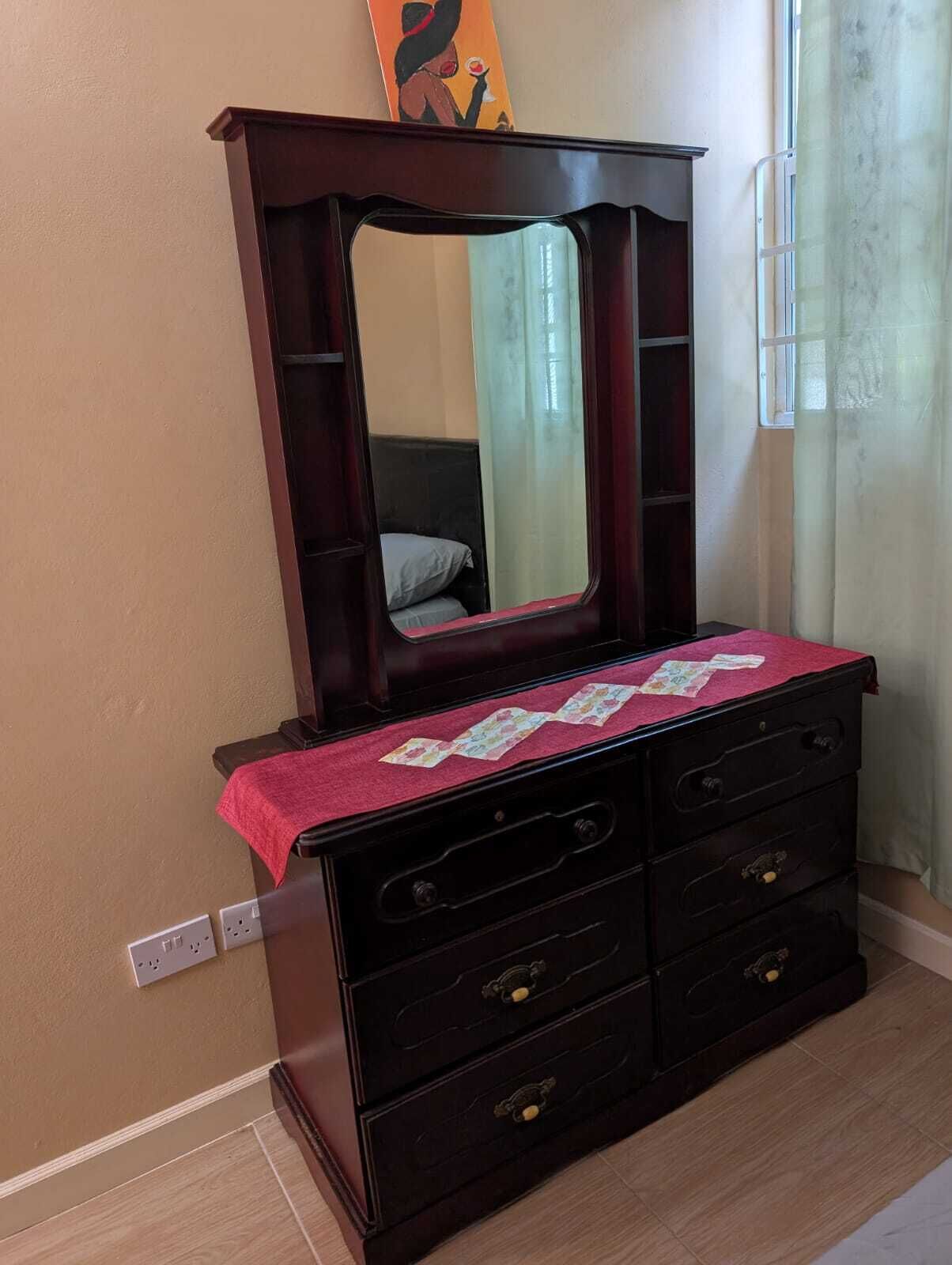 A dark wooden dresser with a mirror above it, a red cloth on top, and a bed in the background.