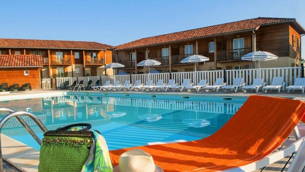 A pool area with lounge chairs and umbrellas, a wooden building with balconies, and a clear blue sky. - Apartment in Eauze With Private Pool (Eauze)