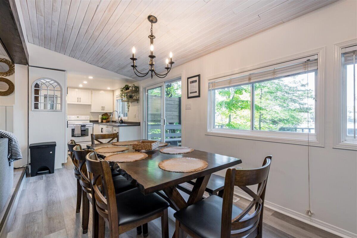 A dining area with a wooden table, chairs, and a chandelier. There is a kitchen in the background with white cabinets and a window.