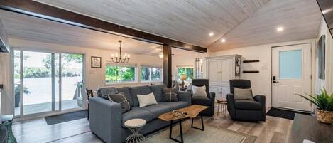 A spacious living area with a grey sofa, a coffee table, and a dark armchair. There is a large window providing a view of the outdoors.
