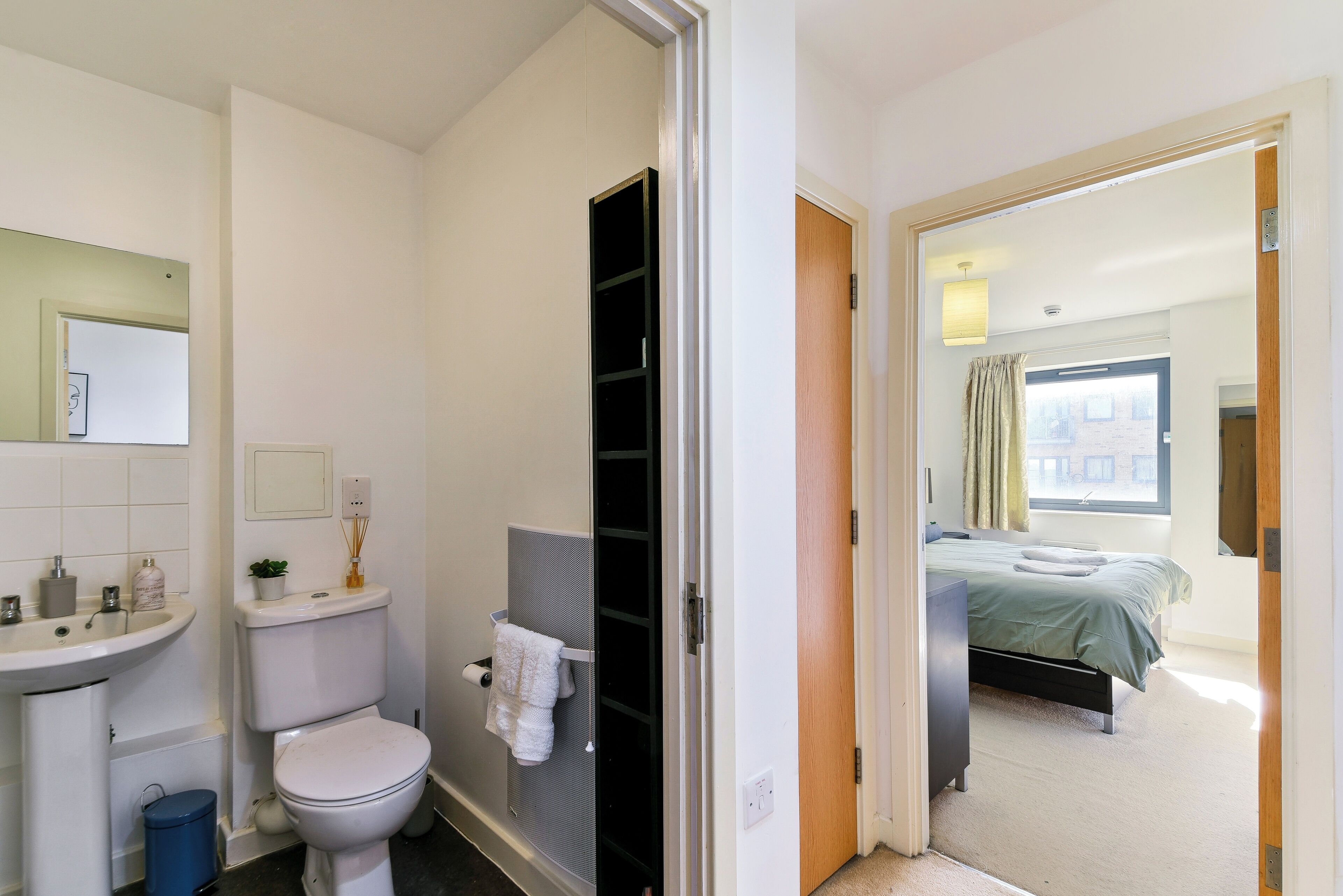 A bathroom with a toilet, sink, and mirror. There is a door leading to a bedroom with a bed and a window.
