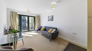A modern living room with a glass dining table, a grey sofa with yellow cushions, a potted plant, and a view of a cityscape through large windows. - Modern 1 bed in Bow - Free Parking (London)