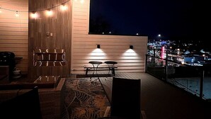 An outdoor seating area with string lights, a wooden wall, and a view of a cityscape at night. - Guest Favorite · Large Lux Rooftop Oasis House  · 4BR · Centrally Located! (Berry Hill)