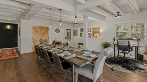 Un comedor con una larga mesa de madera dispuesta para seis personas, rodeada de sillas. A la izquierda, una pizarra, una puerta de madera con un diseño geométrico, y a la derecha, un escritorio con una silla.