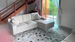 Un salón con un sofá blanco, una escalera de madera y vistas a zonas verdes a través de una ventana con cortinas blancas.