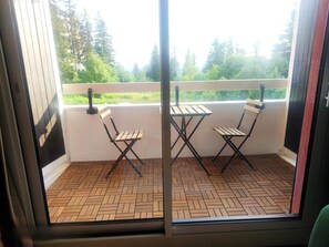 Un balcon avec deux chaises pliantes en bois et une table, un parquet à motifs en chevrons et une vue sur les arbres et la verdure.