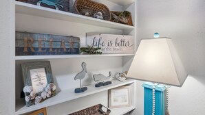 Uno scaffale con oggetti decorativi, tra cui un cartello con la scritta "life is better at the beach", un cartello di benvenuto incorniciato e diverse piccole sculture.