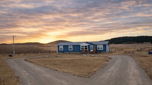 A single-story house with a blue exterior, a front porch, and a dirt driveway leading to the entrance.