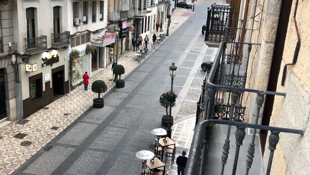 Vista a nivel de calle de un edificio con suelo a cuadros, mesas y sillas para disfrutar al aire libre, y un balcón con barandillas de metal decorativo.