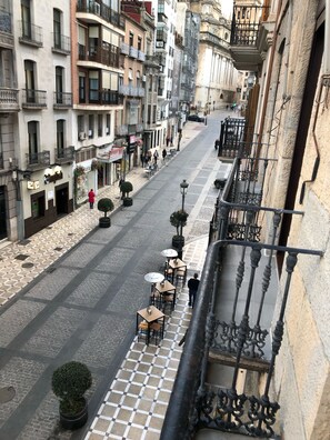 Vista da rua de um edifício com piso em padrão xadrez, área de estar externa com mesas e cadeiras, e uma varanda com grades de metal ornamentadas.