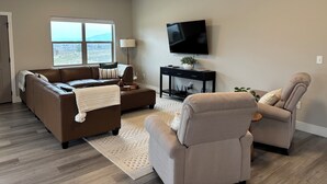 Een woonkamer met een bruine leren bank, een beige fauteuil, een klein bijzettafeltje, een flatscreen televisie aan de muur en een raam met uitzicht op een stadsbeeld.