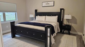 Een slaapkamer met een zwart bedframe, wit beddengoed, twee nachtkastjes met lampen, een raam met zonwering en een bord aan de muur.