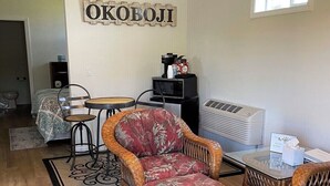 A cozy living room with a wicker sofa, a glass coffee table, and a ceiling fan. A sign with "OKOBOIJI" is on the wall, and there's a window providing natural light.