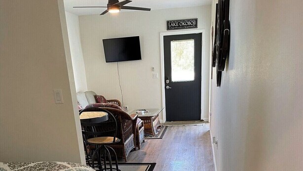 A hallway leading to a bedroom with a bed, a ceiling fan, a TV, and a door labeled "LAKE OKOBOI".