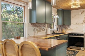 A modern kitchen with dark green cabinets, a wooden countertop, and a large window overlooking trees.