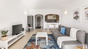 Una sala moderna con un televisor de pantalla plana, un sofá gris, una mesa de centro y una alfombra colorida. Se ve una cocina al fondo con armarios blancos y una zona de comedor con sillas blancas.