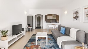 A modern living room with a flat-screen TV, a grey sofa, a coffee table, and a colorful rug. There is a kitchen in the background with white cabinets and a dining area with white chairs. - T1 Moderno  w/Pool in Albufeira by LovelyStay (Albufeira)