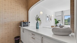 Un baño moderno con tocador blanco, microondas, un florero con plantas y un espejo grande que refleja vistas a un balcón.