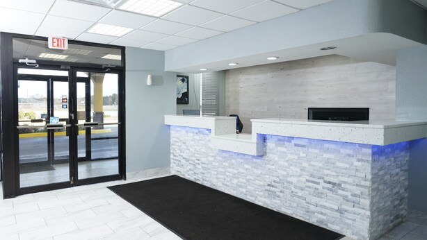 A reception area with a glass door entrance, a reception desk with a stone-like design, and a black doormat. - Red Carpet Newnan (Newnan)