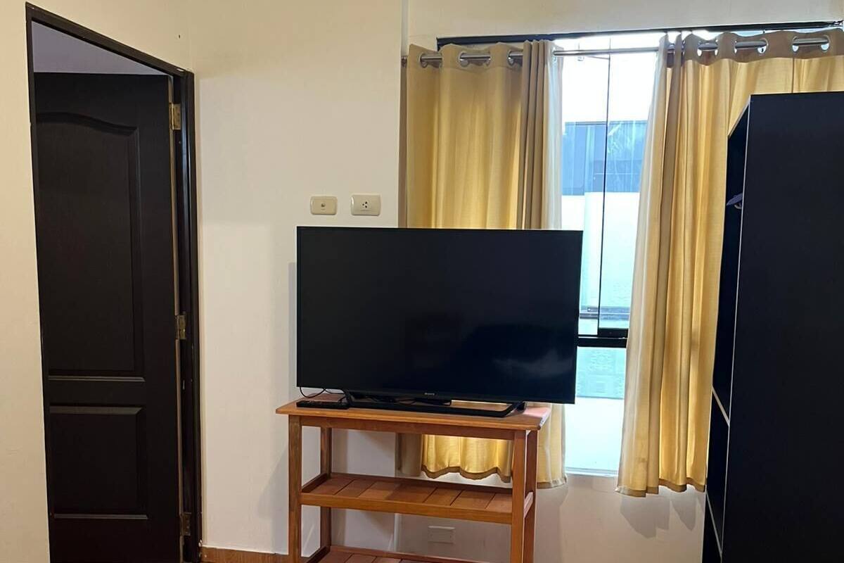 Ένα δωμάτιο ξενοδοχείου με ένα κρεβάτι, μια τηλεόραση τοποθετημένη σε ένα ξύλινο έπιπλο, μια πόρτα και ένα παράθυρο με κουρτίνες.