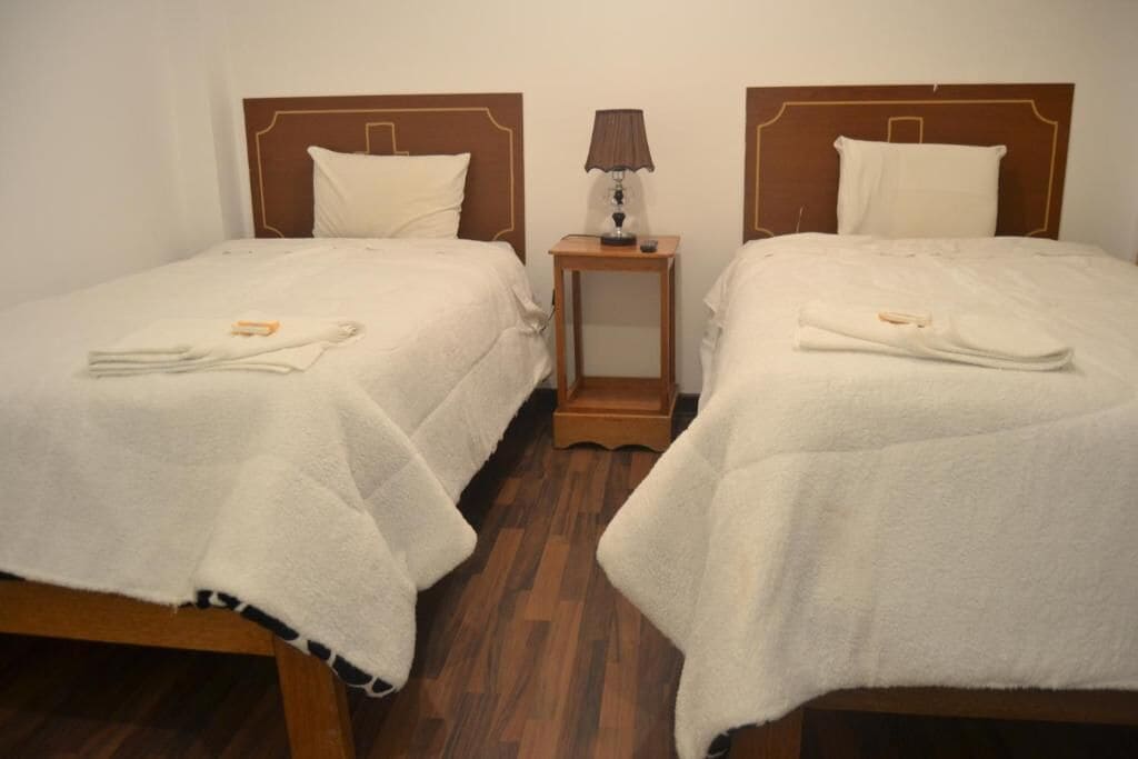 Habitación de hotel con dos camas individuales, cada una con cabecero de madera y ropa de cama blanca. Entre las camas hay una mesita de noche de madera con una lámpara.