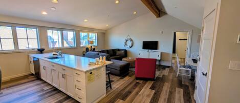 A spacious kitchen with a large island, a dining area with a red chair, a television, and a cozy seating area with a sofa and a decorative wall hanging.