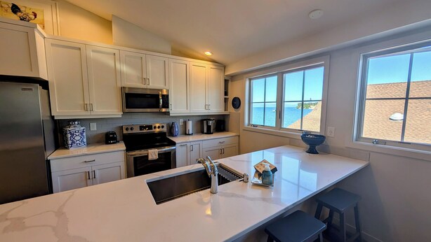 A modern kitchen with white cabinets, stainless steel appliances, a large window with a beach view, and a central island with stools.