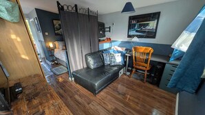 A compact living space with a black leather sofa, a wooden chair, a desk with a computer, and a blue hanging lamp.