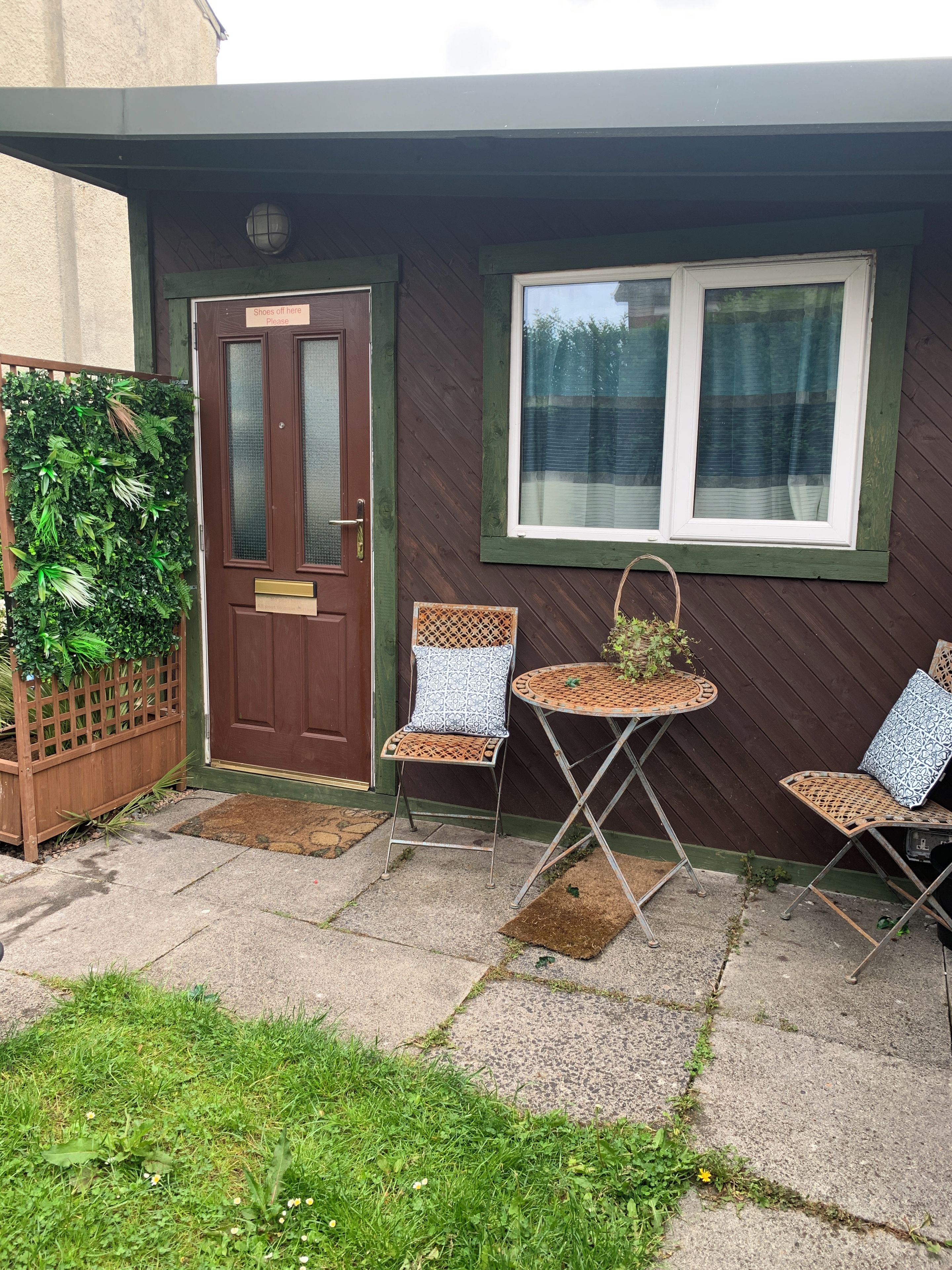 A house with a brown door, a window with a curtain, and two wicker chairs with cushions.
