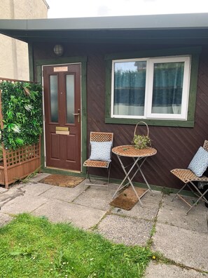 A house with a brown door, a window with a curtain, and two wicker chairs with cushions.