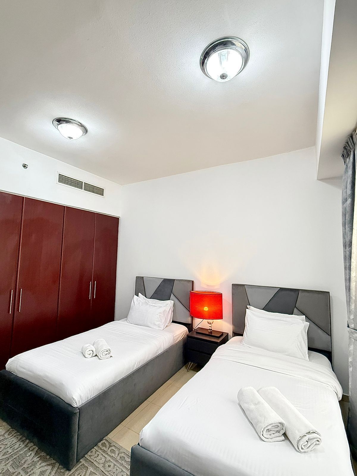 Une chambre d’hôtel avec deux lits simples, une armoire, une table de chevet avec une lampe et un luminaire au plafond.