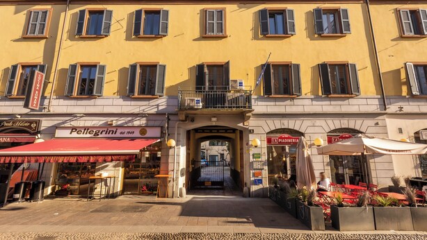 Un edificio giallo con numerose finestre e balconi. È presente un negozio con un tendone rosso e un cartello con su scritto “Pellegrini dal 1949”.