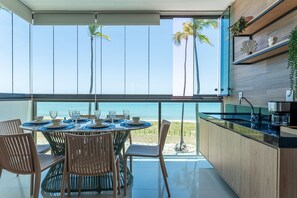 A dining area with a view of the beach, a kitchen with a sink, and wooden cabinets. - 143 - Apto Serrambi Exclusive Living (Ipojuca)
