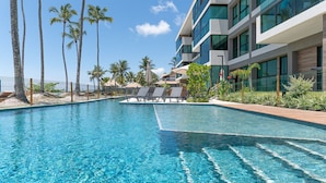 A clear blue pool with lounge chairs and palm trees, surrounded by a modern building with large windows.