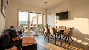 A modern living room with a sofa, a dining area with chairs and a table, a flat-screen TV mounted on the wall, and a sliding glass door leading to an outdoor patio. - Two-bedroom family rooms apartment Casa Del Mar 4 with private balcony (Stara Novalja)