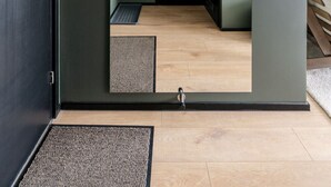Ein Zimmer mit einer dunklen Tür, einer kleinen Eingangshalle mit Fußmatte, einem Holzboden und Blick durch einen Bogen in einen weiteren Raum.
