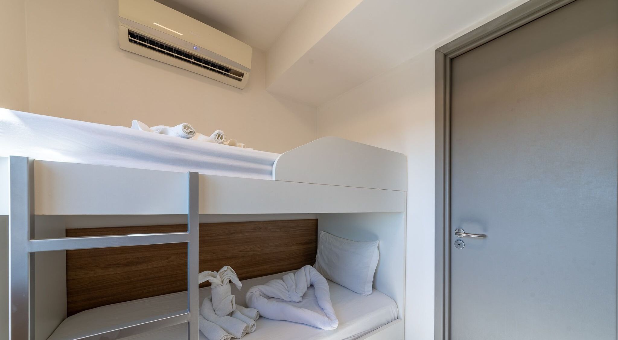 A bunk bed with a wooden headboard, a white mattress, and rolled towels. There is a door to the right and an air conditioning unit above the bed.