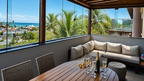 Een kamer met een houten plafond, grote ramen, een bank, een tafel met glazen en een fles, en uitzicht op palmbomen en een strand.