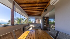 Een balkon met een houten tafel en stoelen, een bank en een glazen wand met uitzicht op de oceaan en palmbomen.